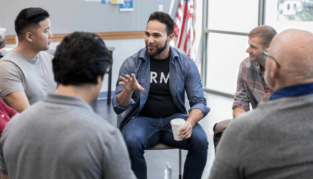 Male veterans talk together in a group setting, smiling, with the American flag in the background.