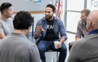 Male veterans talk together in a group setting, smiling, with the American flag in the background.